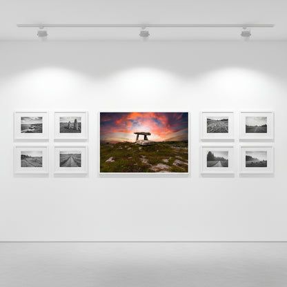 Dolmen print as the focal art piece on gallery-white walls, matte oak frame, spotlights overhead, alongside smaller black-and-white landscape prints.